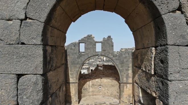 <h2 class="gallery-item-title">ZAMANDA YOLCULUK SUNUYOR</h2>                <p>Tarih boyunca Akad, Amori, Kenan, Arami halklar�n�n yan� s�ra Roma ve Bizans gibi b�y�k uygarl�klar�n etkisi alt�nda kalan Busra'daki tarihi yap�lar, ziyaret�ilerine adeta zamanda yolculuk imkan� sunuyor.<br><br>B�lgede Roma Tiyatrosu (antik tiyatro), Busra Kalesi, Serir Bint el-Melik Tap�na��, Zafer Kemeri, El-Hawa ve El-Nabati kap�lar� gibi �ne ��kan tarihi yap�lar yer almaktad�r.<br><br>Busra'n�n simge yap�lar�ndan Roma Tiyatrosu, �mparator Trajan d�neminde 2. y�zy�lda in�a ettirildi. 15 bin ki�ilik kapasitesi ve bazalt ta� i��ili�iyle b�lgenin en g�rkemli yap�s� olmay� s�rd�r�yor.<br><br>7. y�zy�ldan itibaren tiyatronun �evresine kal�n sur duvarlar�, kuleler ve bur�lar eklenerek kaleye d�n��t�r�ld�. Yap� ha�l� seferleri s�ras�nda savunma ama�l� kullan�ld�.<br><br>D�nyada tamamen sa�lam kalm�� nadir Roma antik tiyatrolar�ndan biri say�lan yap�y� �evreleyen surlar ve kuleler hala g�r�lebiliyor.<br><br>Suriye'deki i� sava�ta antik kentteki bir�ok tarihi yap� devrik rejim g��lerinin sald�r�lar� sonucu zarar g�rd�.<br><br>Turizmin yeniden canlanmas� durumunda Busra, yeniden turizm merkezlerinden biri olabilir.</p>